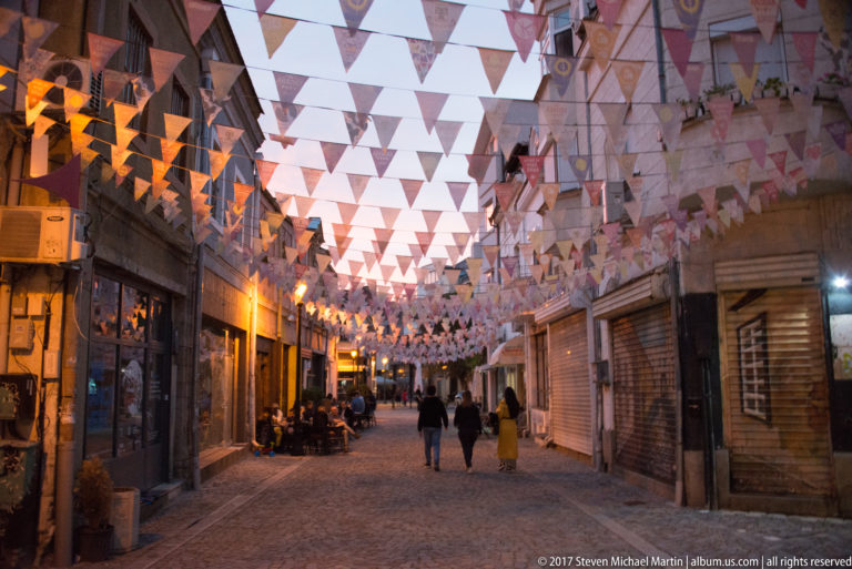 People walking beneath rows of triangular bunting on a warmly lit cobbled pedestrian street at dusk.