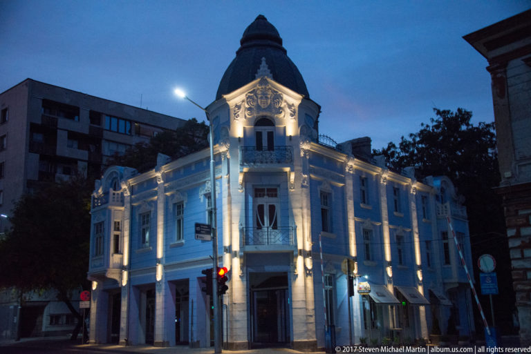 Illuminated white historic corner building with ornate façade, projecting bay, and rounded dark roof at blue hour.