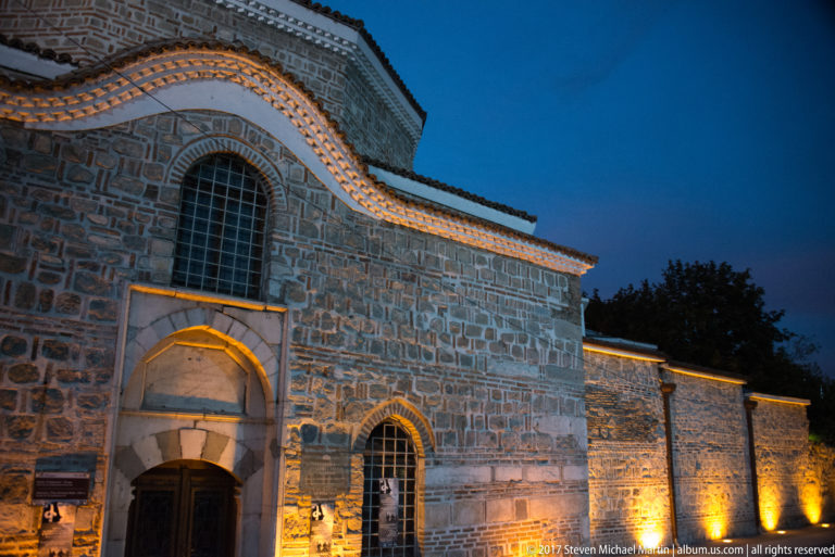 Illuminated stone church with arched doorway, tall window, gabled roofline, and long side wall at blue hour.