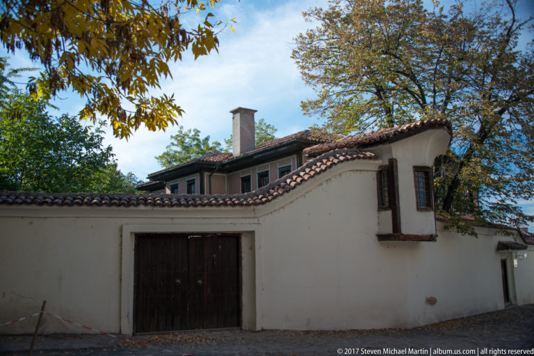 Whitewashed courtyard wall with wooden gate, red tile coping, and traditional upper-story windows beneath leafy trees.