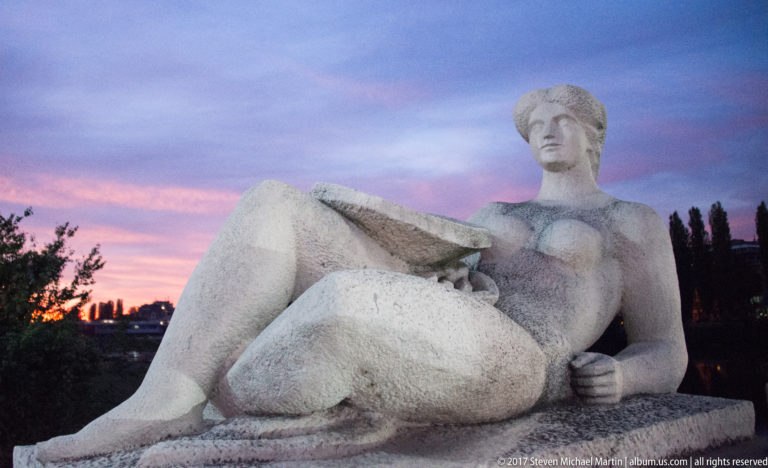 Large pale stone sculpture of a reclining woman with one knee raised against a pink-and-blue evening sky.
