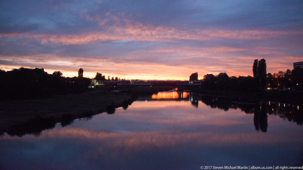 Calm Maritsa River reflecting a pink-orange sunset, dark tree silhouettes, and a distant illuminated bridge in Plovdiv.