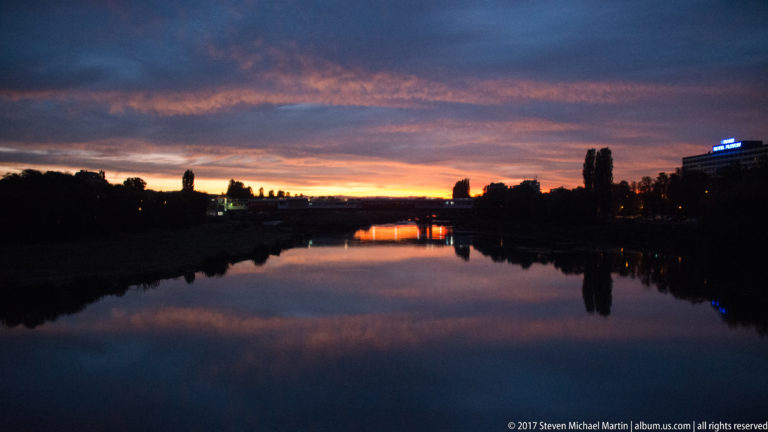 Darkening Maritsa River reflecting blue-violet clouds and a narrow orange sunset band, with distant bridge and city lights.