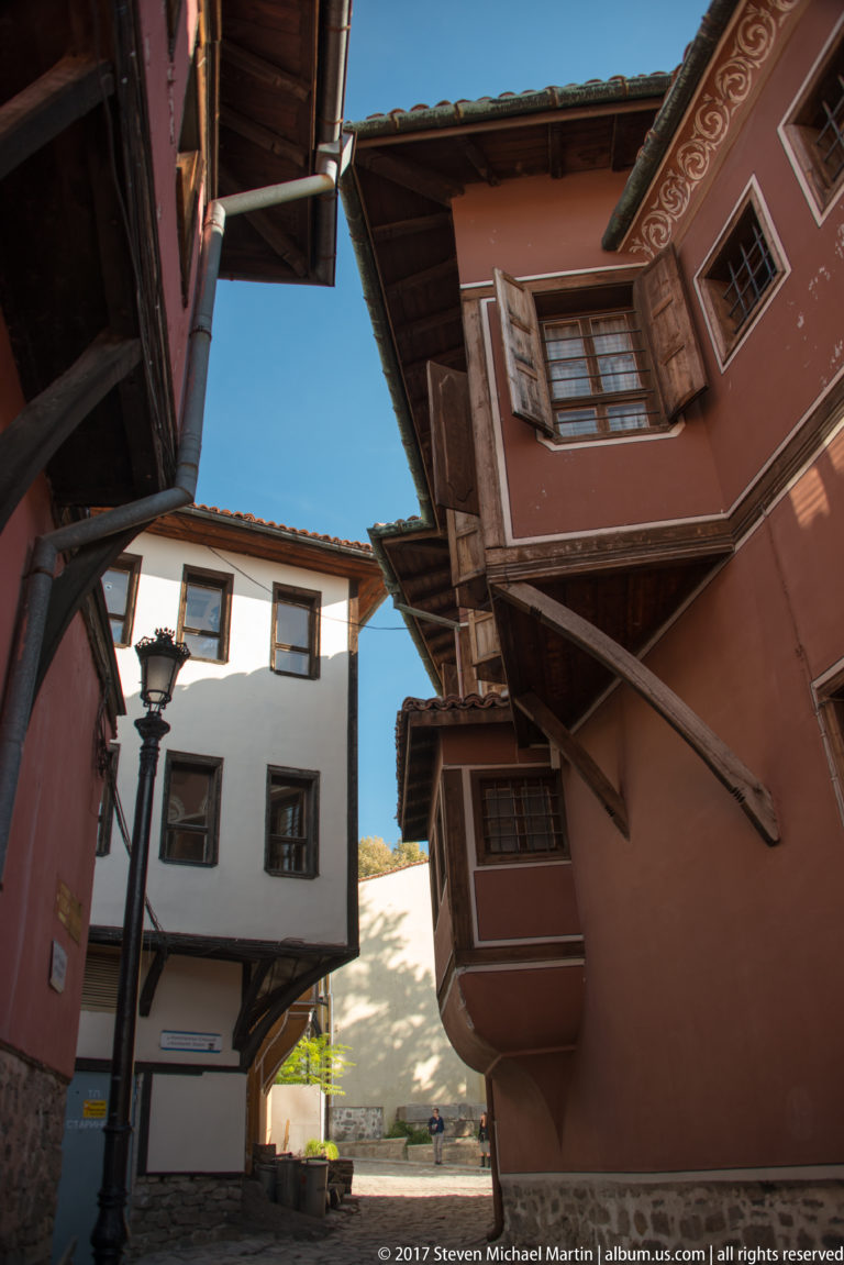 Upward view between red and white traditional houses with projecting upper stories, shutters, deep eaves, and a streetlamp.