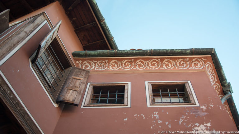 Rose-colored historic façade with white scrolling cornice decoration, wooden shutters, barred windows, and deep tiled eaves.