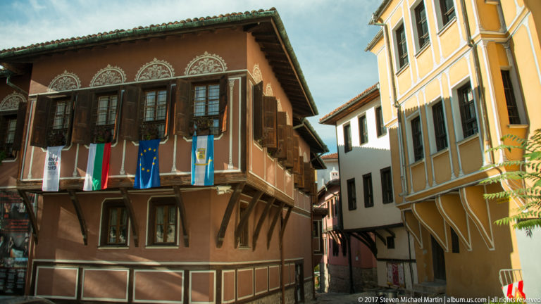 Traditional brown timber-fronted house with rows of shutters and Bulgarian and European Union flags beside a yellow historic building.