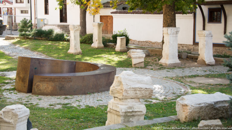 Stone columns, capitals, and carved fragments arranged in a grassy courtyard around a curved rust-colored interpretive installation.