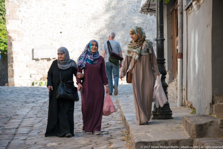Three women in headscarves and long garments walking down a cobblestone lane beside white walls and a black streetlamp.