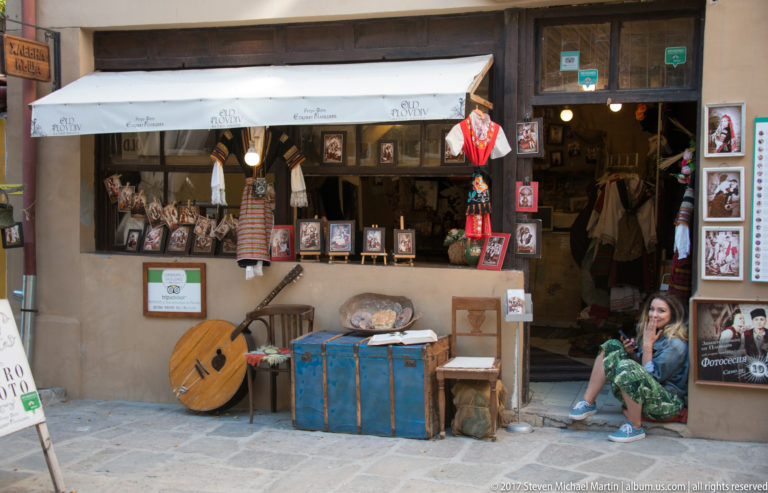 Open-front Old Plovdiv shop filled with framed pictures, textiles, crafts, a stringed instrument, blue trunk, and seated woman.