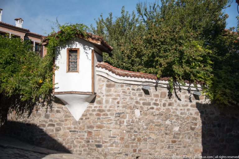 White projecting room with wooden window above a rough stone wall, red tile roof, trees, and shaded lane.