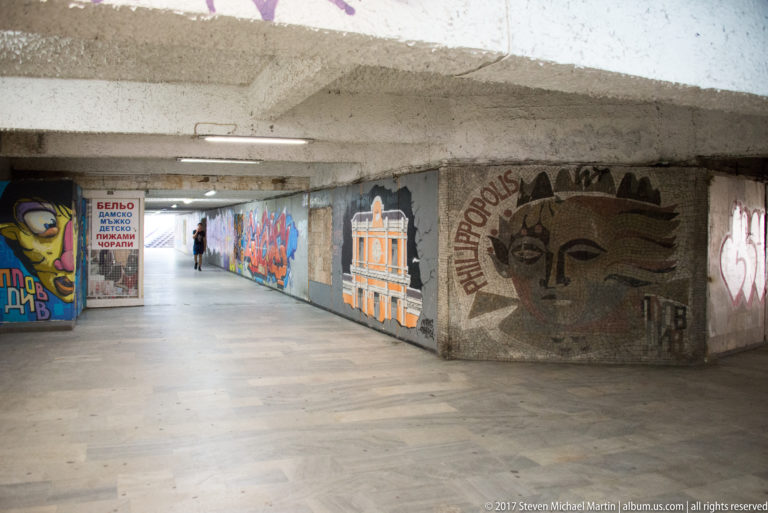 Concrete pedestrian underpass lined with colorful graffiti murals and large painted faces, with daylight visible at the far end.