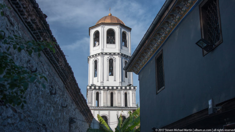 White multi-level bell tower with arched openings and copper-colored roof framed tightly between historic walls.