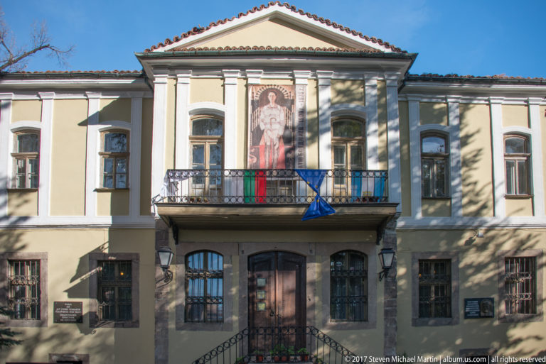 Cream and gray historic building with pediment, pilasters, central balcony, religious icon banner, and Bulgarian and EU flags.