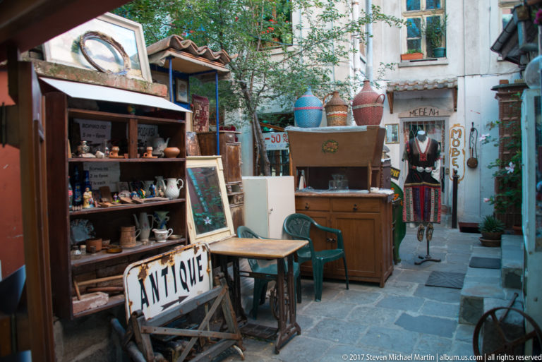 Narrow stone-paved passage with antique sign, pottery, shelves, furniture, clothing, and a mannequin outside small shops.