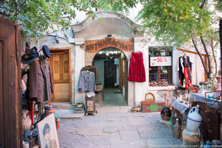 Small arched shop entrance surrounded by hanging clothing, pottery, framed art, and a red “-50%” sale sign.