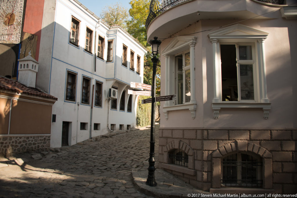 Cobblestone lane between white historic houses with stone foundations, wooden windows, rounded corner bay, and black streetlamp.