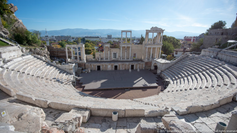 Wide view from upper marble seating across the Ancient Theatre of Philippopolis, stage façade, city, and distant mountains.