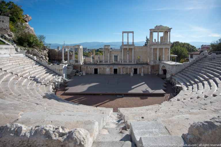 Centered view down pale marble seating toward the stage and columned façade of the Ancient Theatre of Philippopolis.