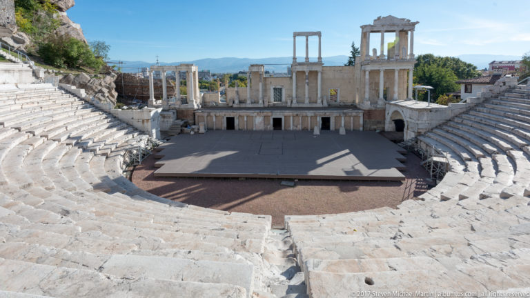 High central view over weathered marble seats toward the Ancient Theatre of Philippopolis stage façade in strong sunlight.
