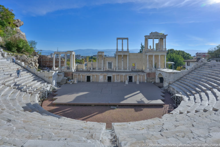 Wide sunlit view of the Ancient Theatre of Philippopolis with marble seating, stage façade, blue sky, hills, and a few visitors.