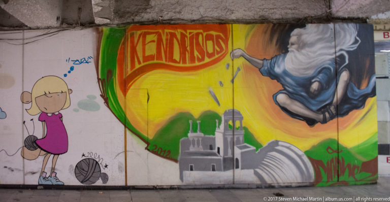 Colorful underpass mural with cartoon figure, large lettering, architectural silhouette, and part of a painted human figure.