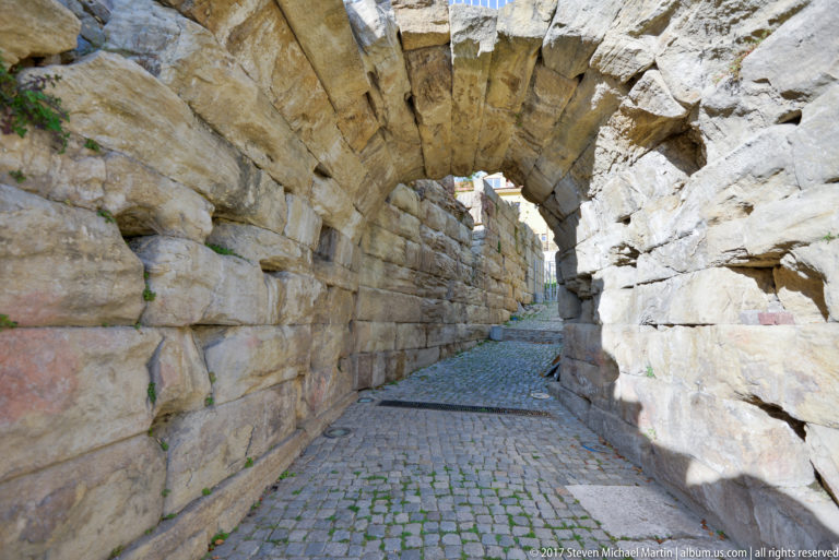 Barrel-vaulted stone corridor beneath the Ancient Theatre of Philippopolis, with block walls and cobblestone paving leading toward daylight.