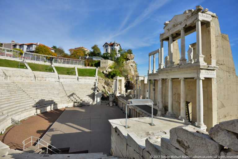 Columned Roman stage façade beside curving stone seating, with houses and greenery above the ancient theatre.