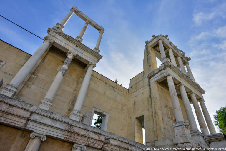 Low-angle view of tall columns, fragmentary entablatures, and pale masonry in the ancient theatre’s stage building.