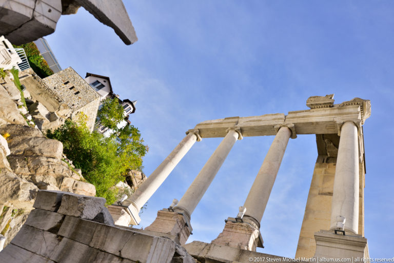 Four pale columns and a fragmentary entablature seen steeply from below against blue sky and rocky masonry.