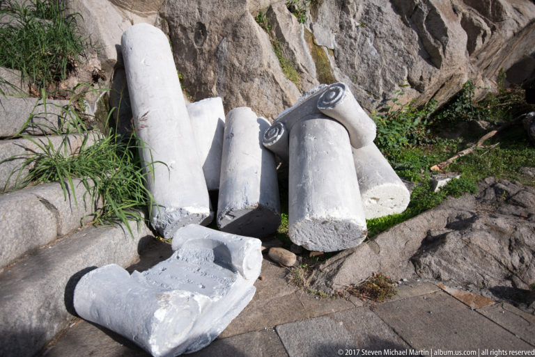 Broken pale stone column drums and a carved capital fragment lying beside rock and grass.