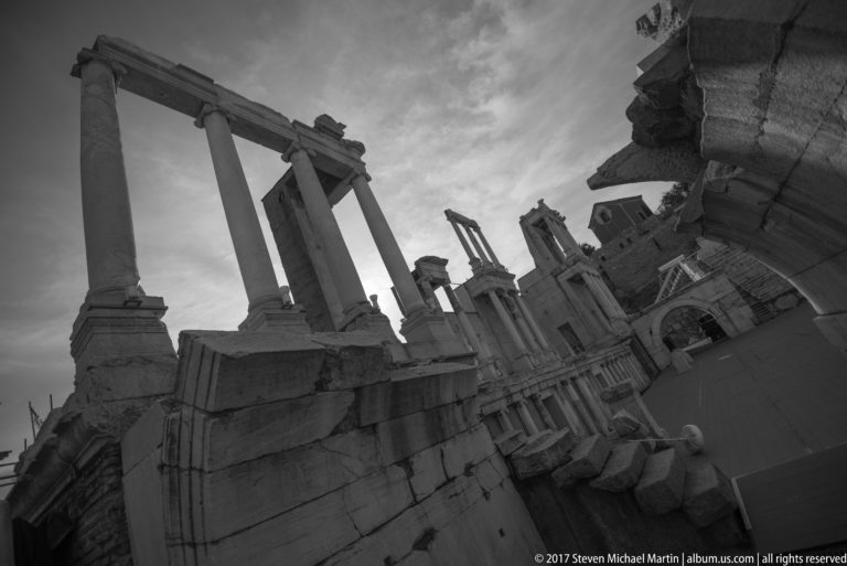 Tilted black-and-white view along columns, masonry, and fragmentary stage architecture at Plovdiv’s ancient theatre.