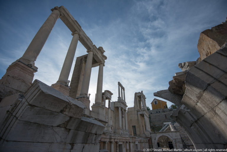 Low view through Roman columns and broken masonry toward the stage structures and a hillside building beyond.