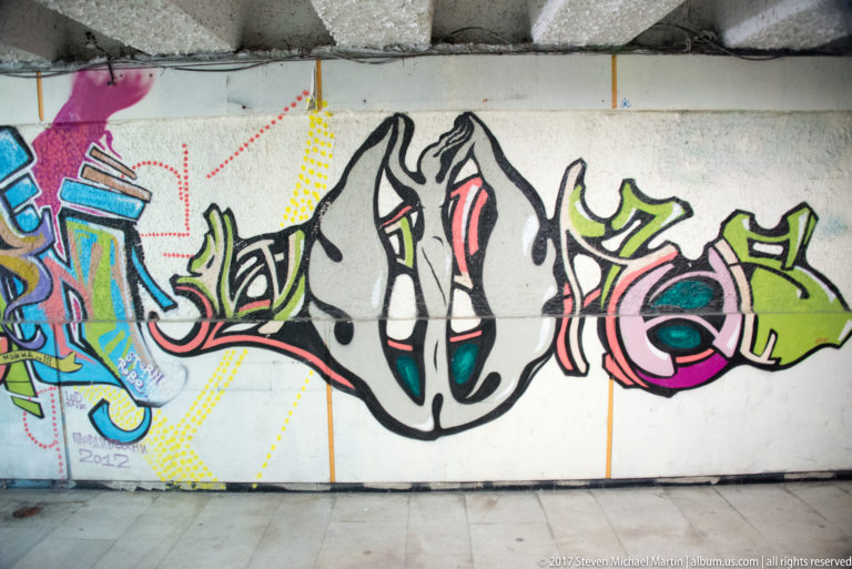 Large black, white, pink, and green graffiti lettering across a tiled wall, with smaller painted forms at left.