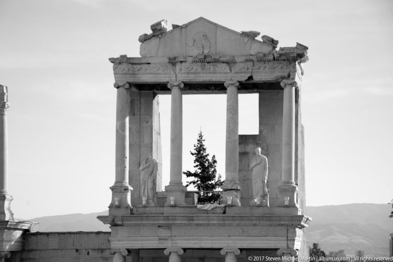 Black-and-white frontal view of a four-column Roman stage structure with two statues and a tree visible between columns.