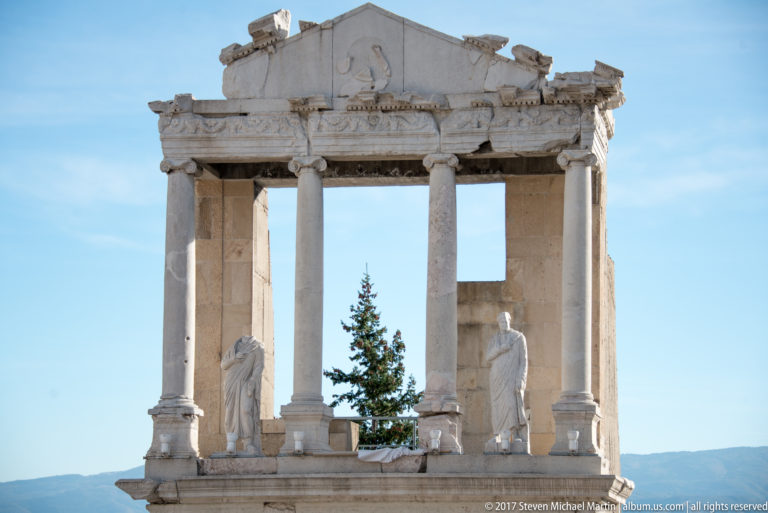 Sunlit four-column stage structure with two draped statues, an evergreen tree, and distant hills visible through the openings.