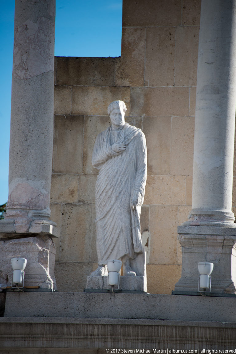 Headless pale stone statue in deeply folded drapery standing between columns against a block-masonry wall.