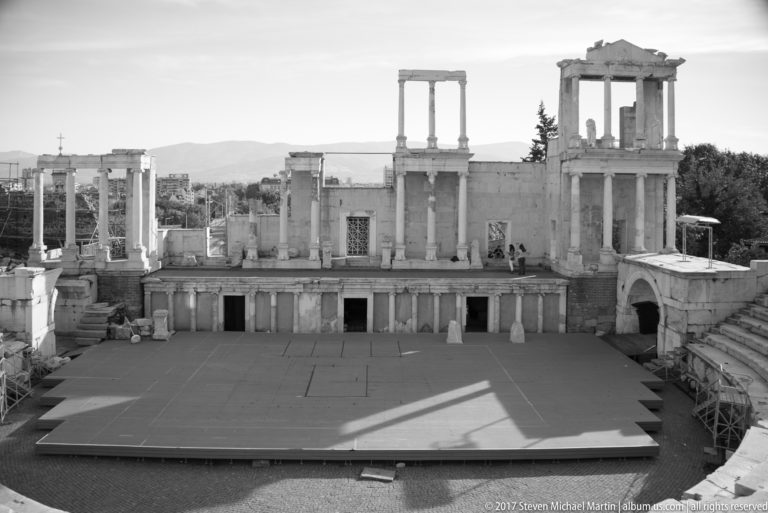 Centered view across the modern stage toward the columned Roman stage architecture, with distant hills beyond.