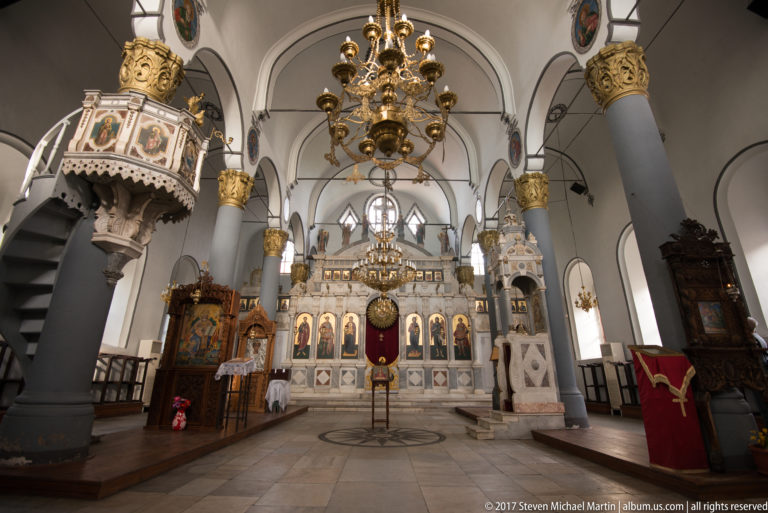 Orthodox church nave with gray columns, brass chandeliers, carved furnishings, and a white iconostasis filled with icons.