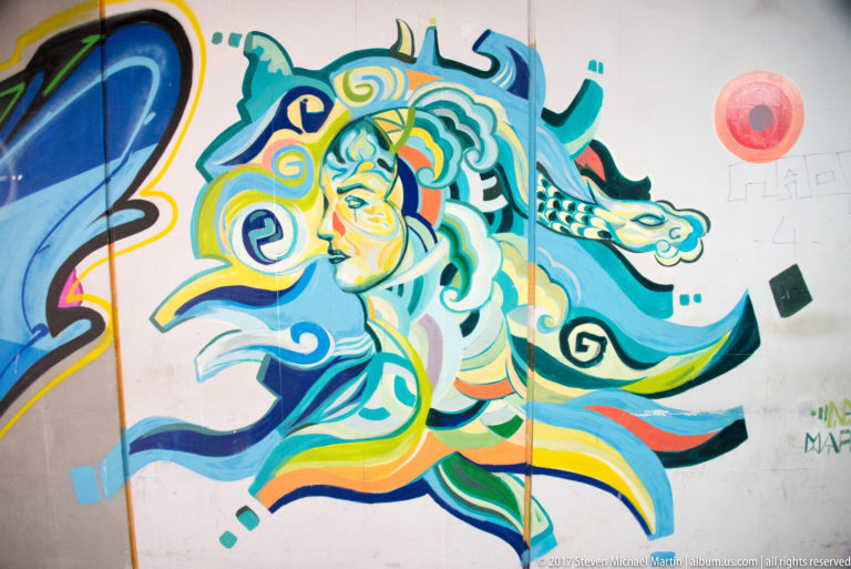Stylized face surrounded by swirling blue, turquoise, yellow, and orange forms with a dragon-like figure on a white wall.