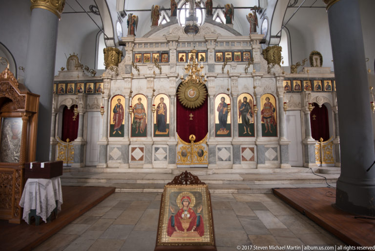 White Orthodox iconostasis with rows of painted saints, gilded details, red curtains, and a framed icon in front.