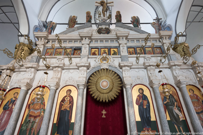 Upper white iconostasis with saint icons, gilded ornament, radiant circular motif, red curtain, and crucifixion figures above.