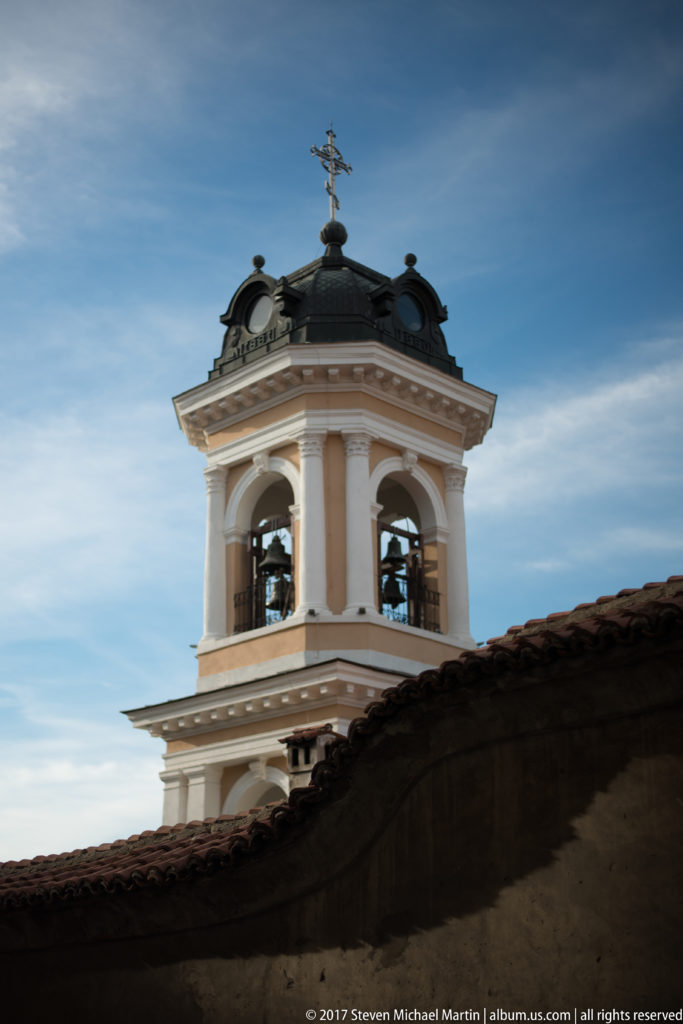Warm-toned church bell tower with open arches, visible bells, dark roof, and Orthodox cross above a tiled roof.