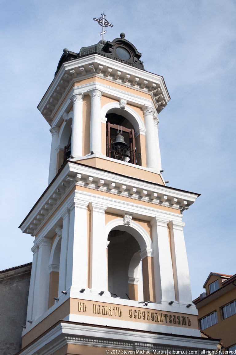 Close view of a pale Orthodox bell tower with arched openings, bells, white trim, cross, and Bulgarian inscription.