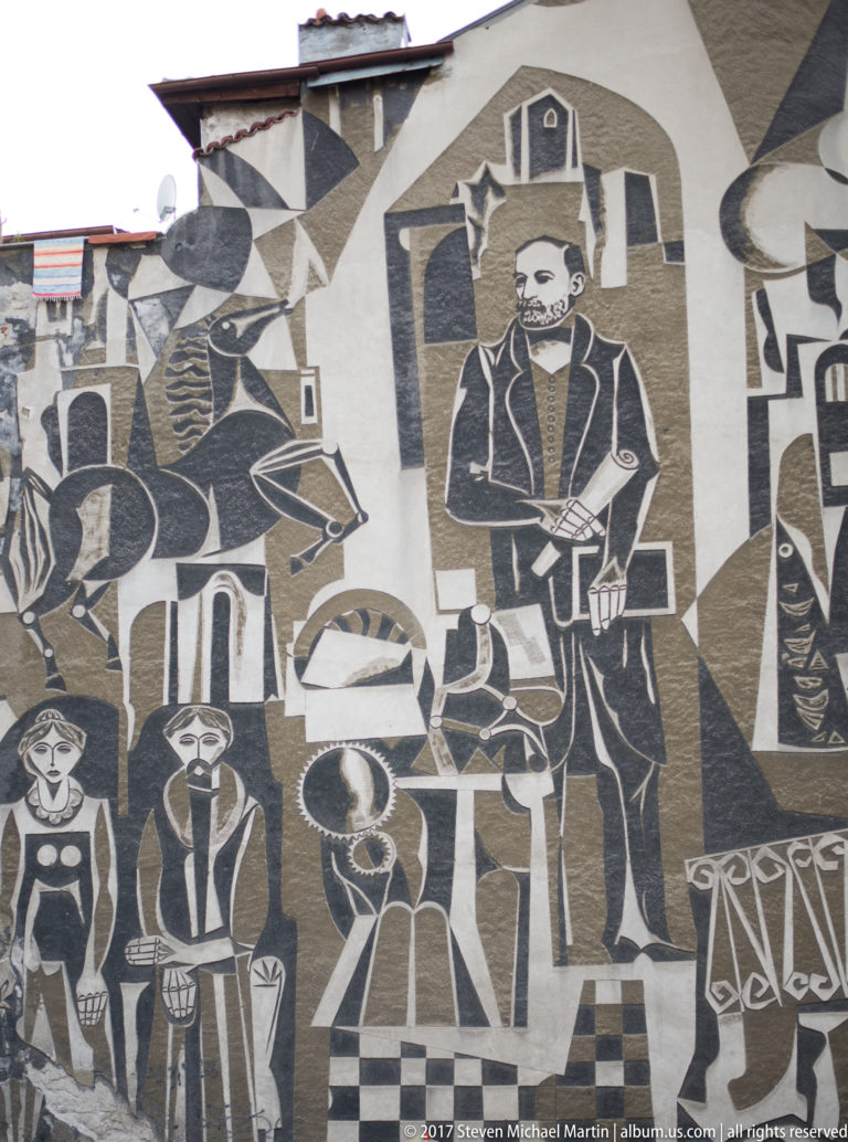 Large black-and-white wall mural with a central bearded man, smaller figures, vessels, buildings, and geometric patterns.