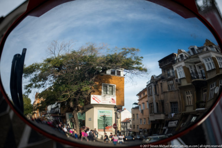 Convex mirror reflecting pedestrians, trees, storefronts, and multi-story buildings along a street in Plovdiv.