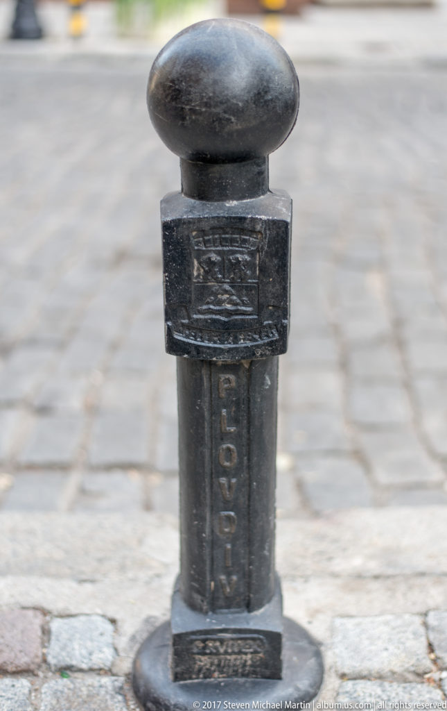 Dark cast-metal bollard with rounded cap, square collar, and raised vertical markings on pale cobbled paving.