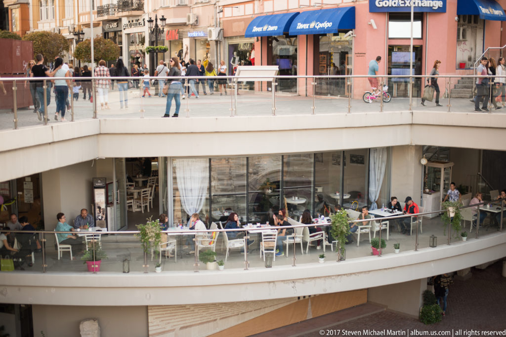 People dining on lower café terraces while pedestrians walk past storefronts on an elevated level above.