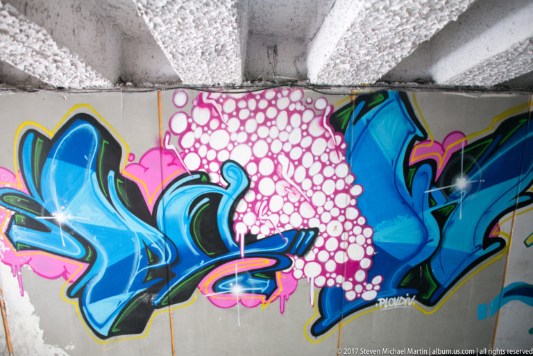 Blue graffiti lettering with black outlines beside a dense pink bubble pattern beneath a concrete underpass ceiling.