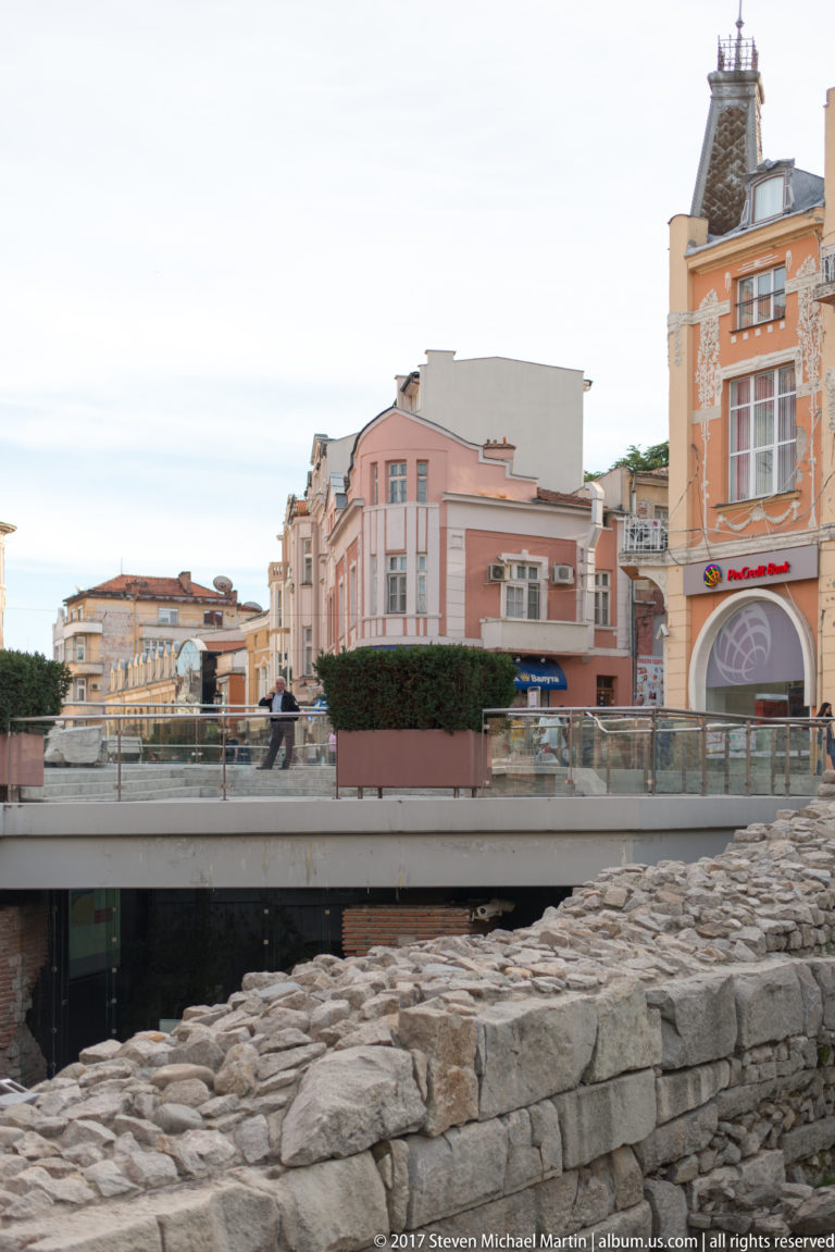 Low ancient stone walls in the foreground with a modern pedestrian structure and colorful central Plovdiv buildings beyond.