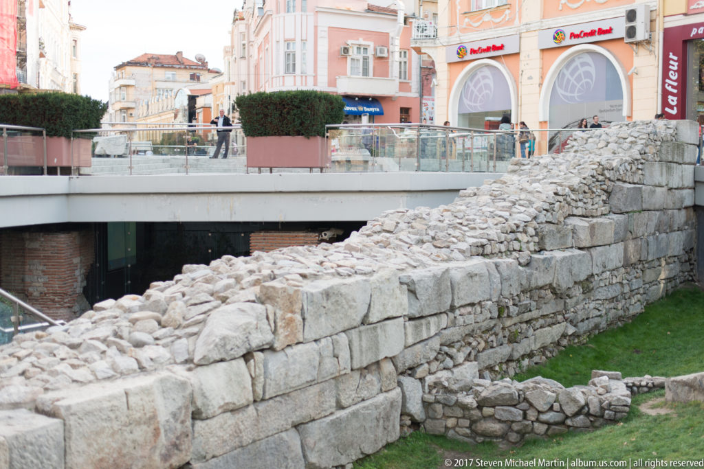 Pale ancient stone walls and rubble rise below a modern pedestrian terrace with shops and people in central Plovdiv.